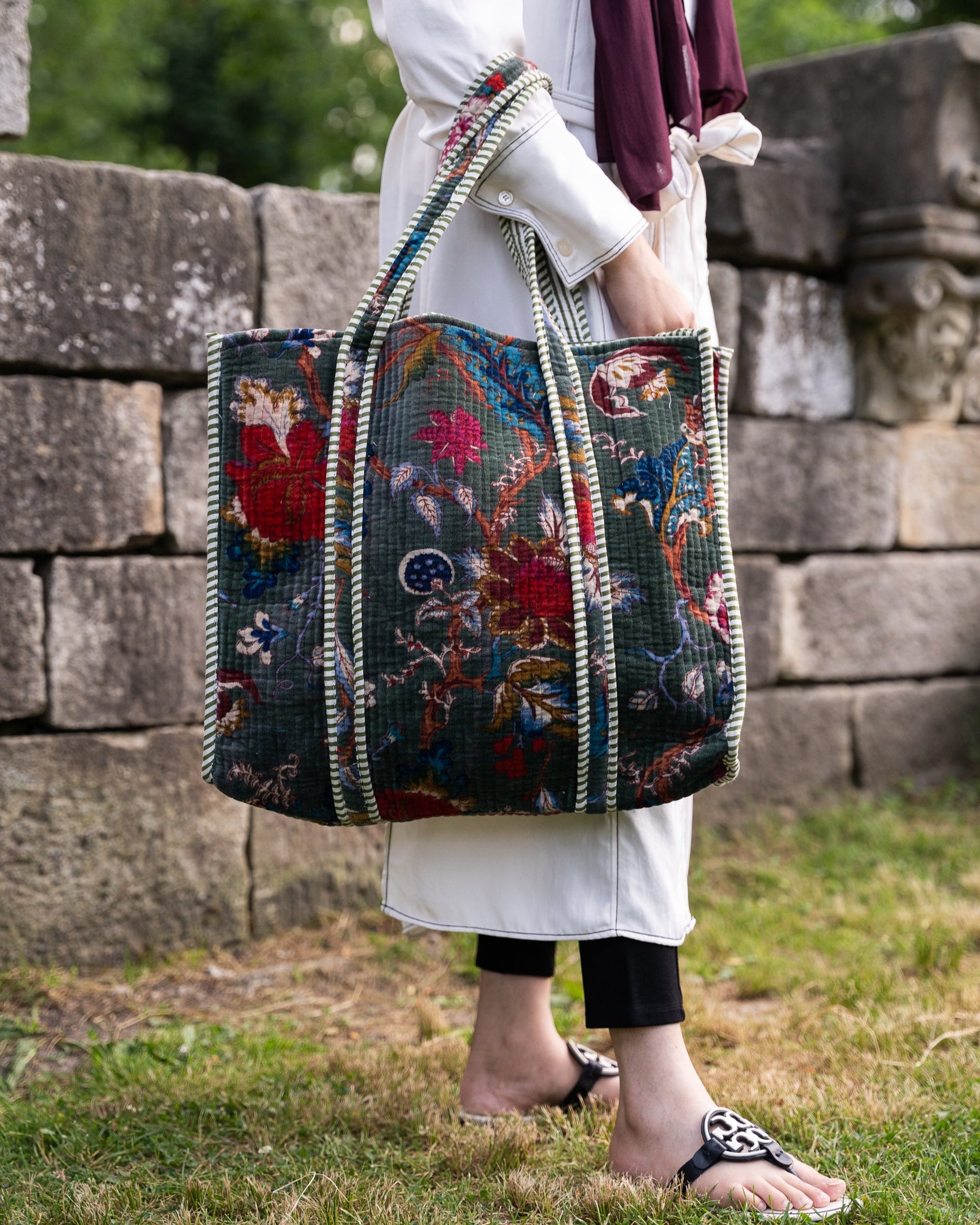 Person holding a colorful embroidered bag outdoors with stone wall and greenery in the background
